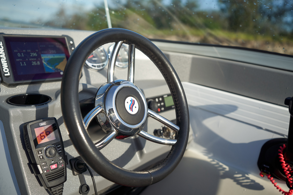 A Quintrex boat's helm featuring a chrome steering wheel, Lowrance navigation screen, and a VHF radio handset.