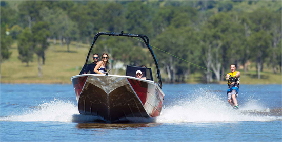 A red and silver Quintrex Freestyler boat towing a water skier across a lake, with passengers on board.