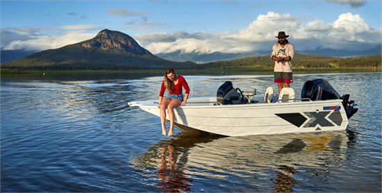 A man and woman on a Quintrex Hornet boat on a calm lake, with a large mountain and cloudy sky in the background.